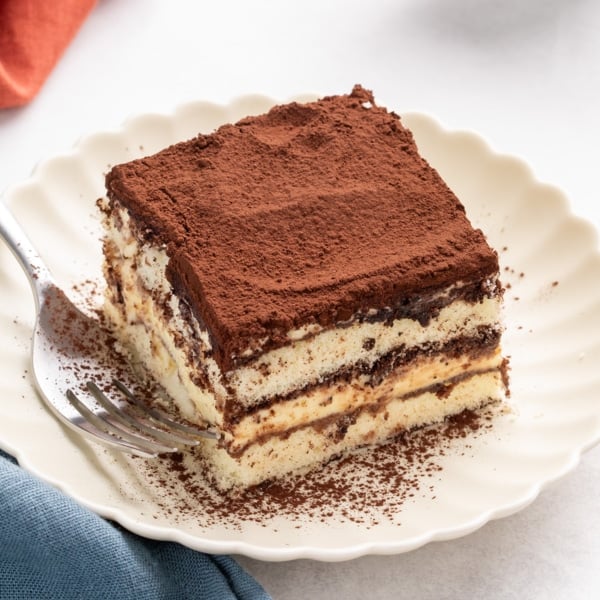Square slice of gluten free tiramisu on a scalloped white plate with cocoa powder on top and a fork beside it.