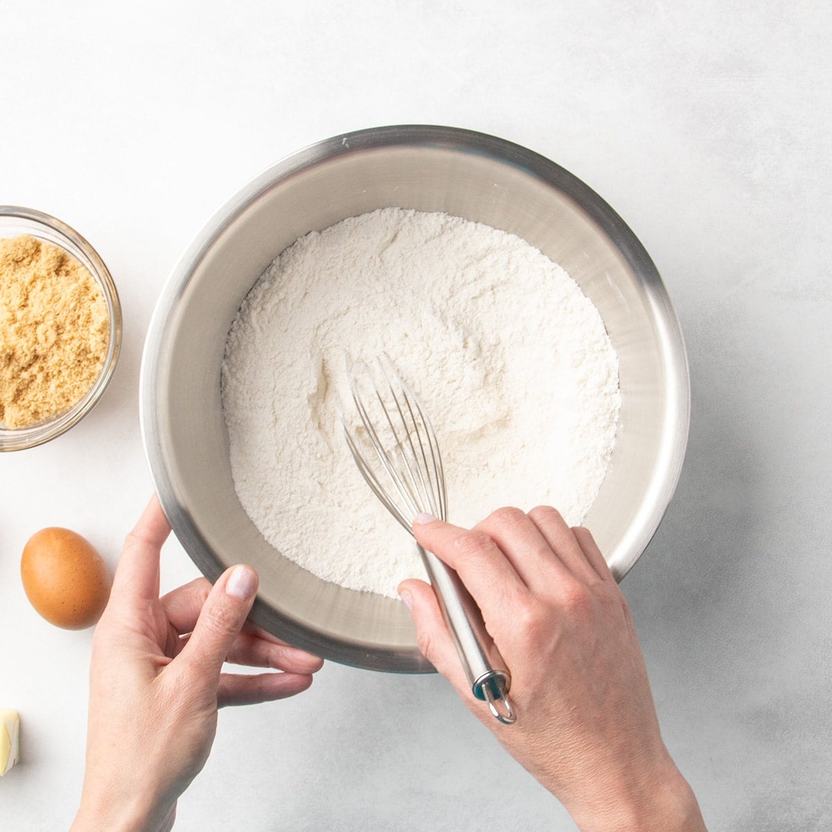 Hands whisking white dry ingredients in a metal mixing bowl with a small bowl of brown sugar and a whole egg nearby.