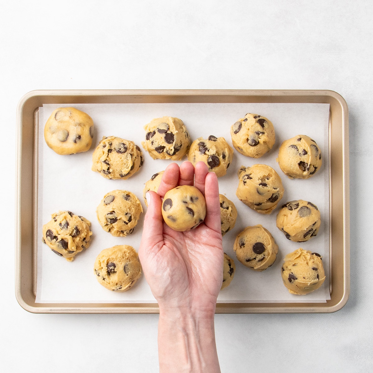 Hand holding a smooth ball of raw chocolate chip cookie dough above a parchment-lined baking sheet with more dough portions in the background.