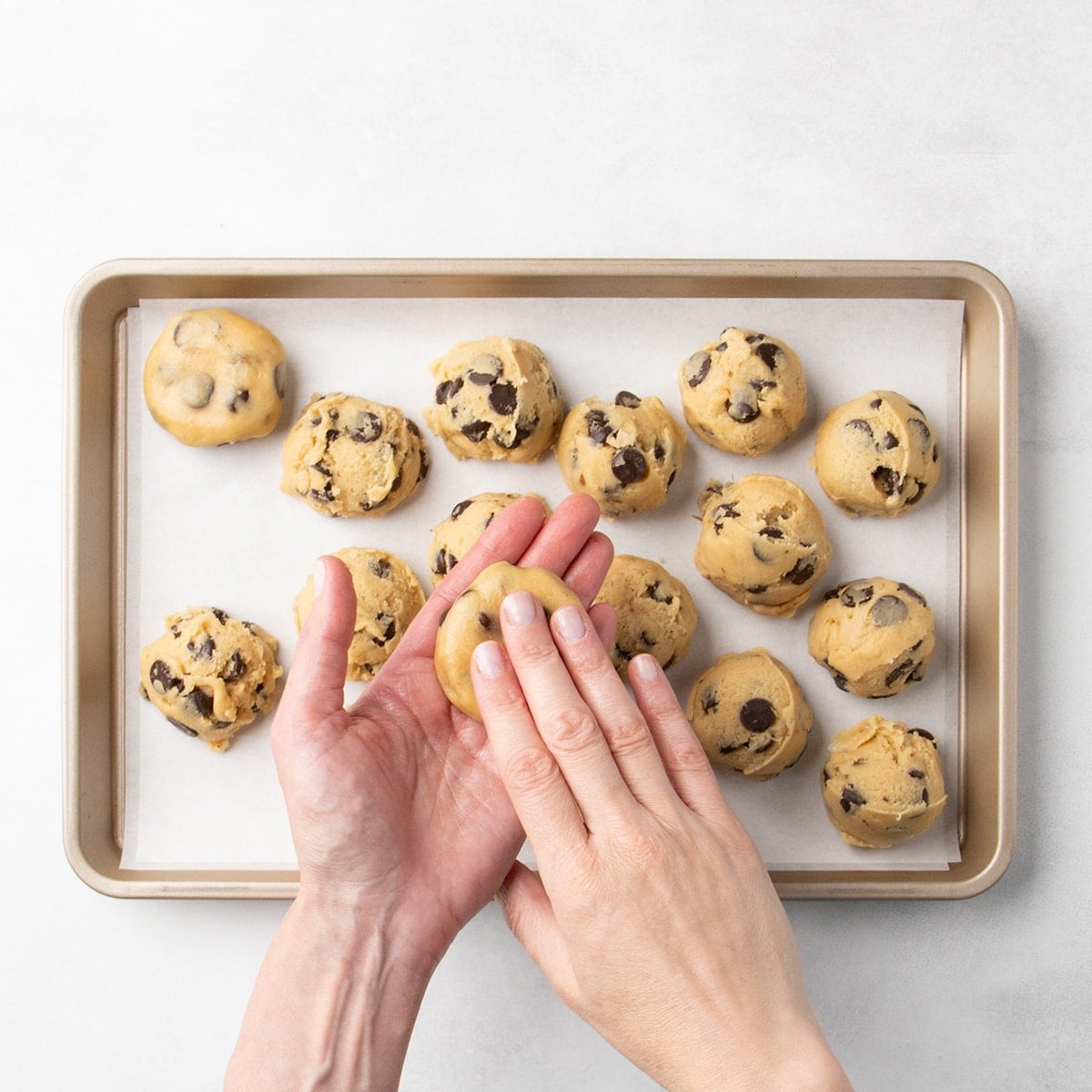 Fingers of one hand flattening a ball of chocolate chip cookie dough in another hand, above a parchment-lined baking sheet with more dough portions in the background.
