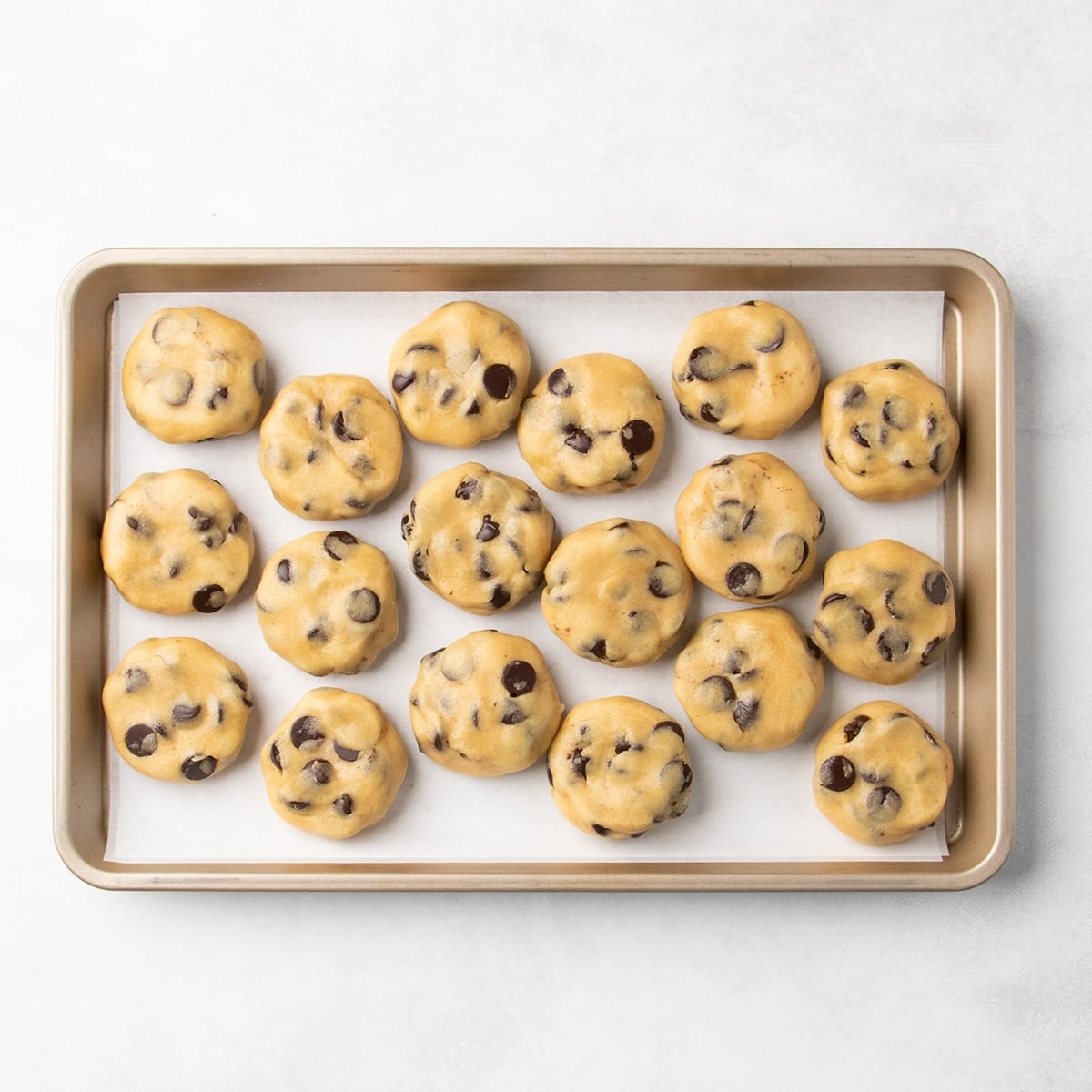 18 flattened raw chocolate chip cookie dough disks arranged on a parchment-lined baking sheet.