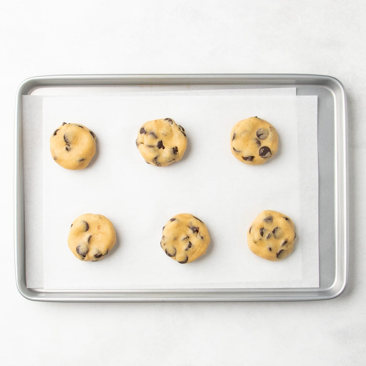 Six flattened chocolate chip cookie dough portions spaced apart on a parchment-lined baking sheet.