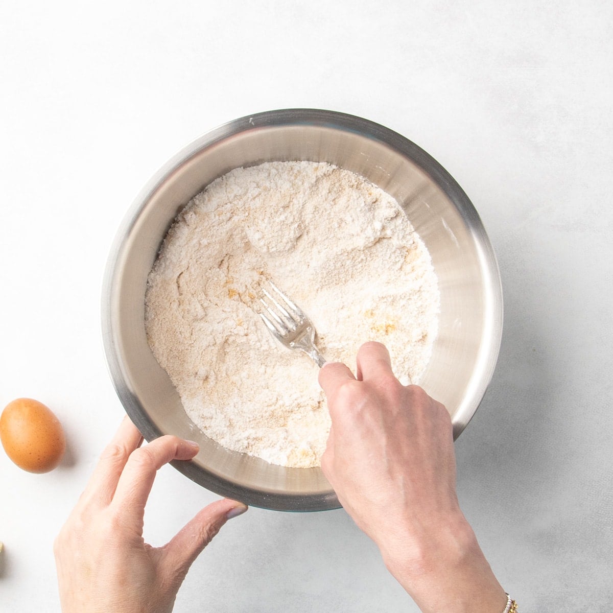 Hands using a fork to mix brown sugar into white flour dry ingredients in a metal bowl.