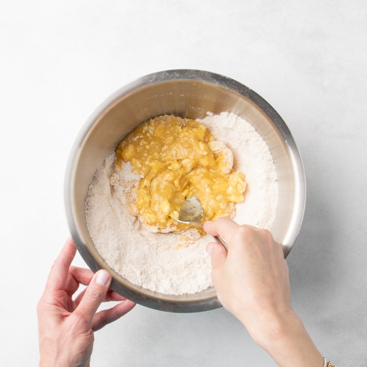 Hand using a large mixing spoon to stir partially mixed cookie dough in a metal bowl with a spoon.