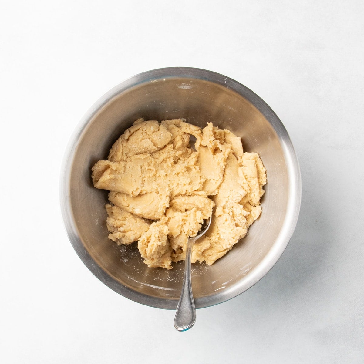 Thick pale brown raw cookie dough in a metal mixing bowl with a spoon resting inside.