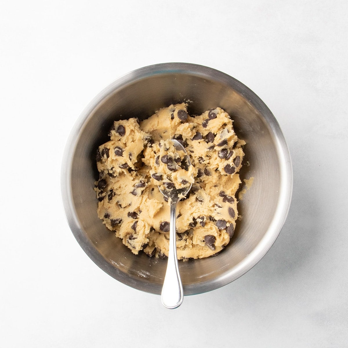 Raw, unshaped chocolate chip cookie dough in a metal bowl with a spoon resting inside.