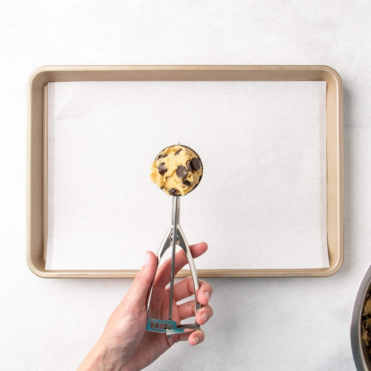 Hand holding a spring-loaded cookie scoop with chocolate chip cookie dough above a parchment-lined baking sheet.