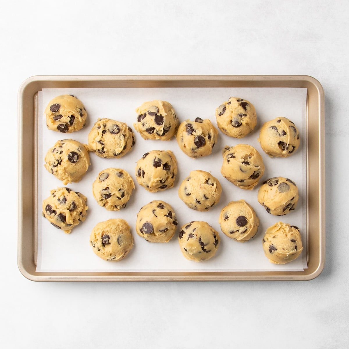 18 mounds of raw unshaped chocolate chip cookie dough arranged on a parchment-lined baking sheet.
