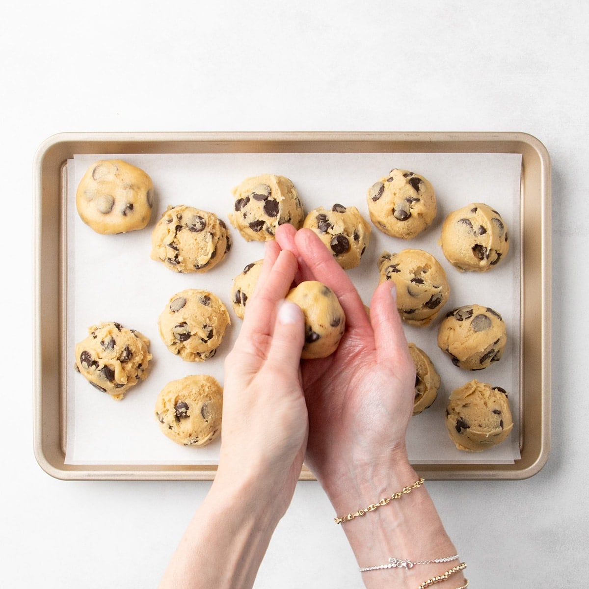 Hands rolling a scoop of raw chocolate chip cookie dough into a ball above a parchment-lined baking sheet with more mounds of raw cookie dough on it.