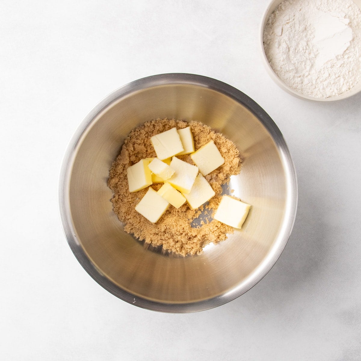 Brown sugar and pieces of butter in a metal mixing bowl with a bowl of white flour nearby.
