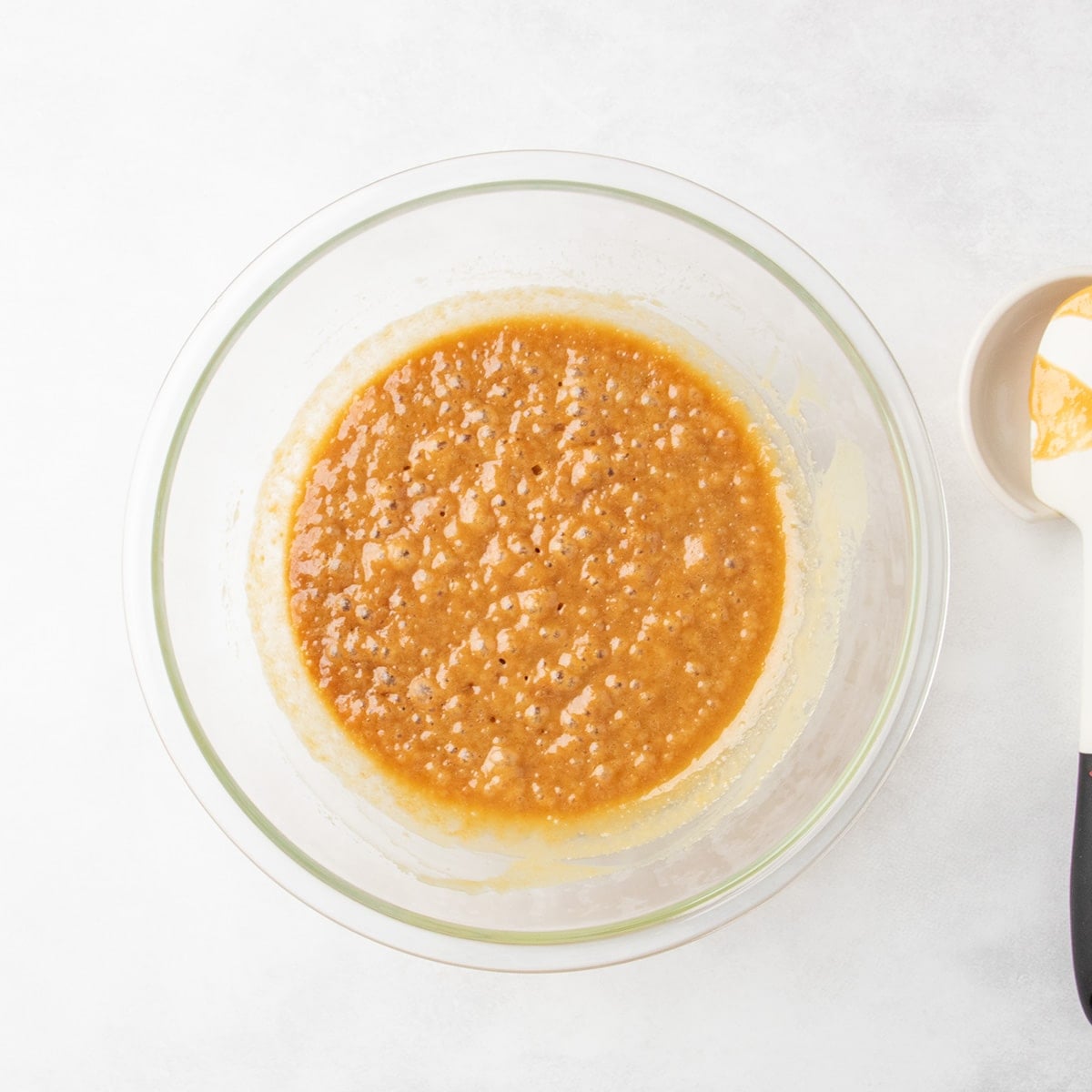Bubbly darker caramel-colored mixture in a clear glass mixing bowl with a spatula nearby.