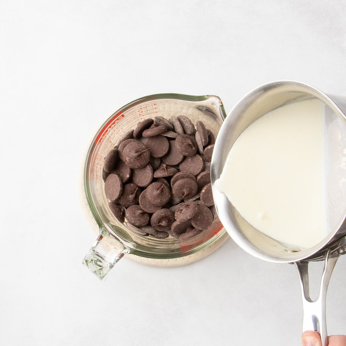 Cream being poured from a small saucepan into a glass measuring cup filled with chocolate pieces.