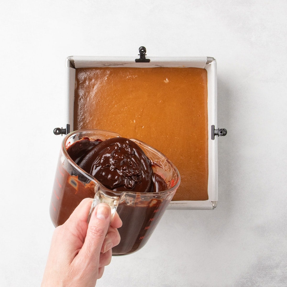 A hand holding a glass measuring cup filled with melted chocolate about to pour it over a pan with a caramel layer.