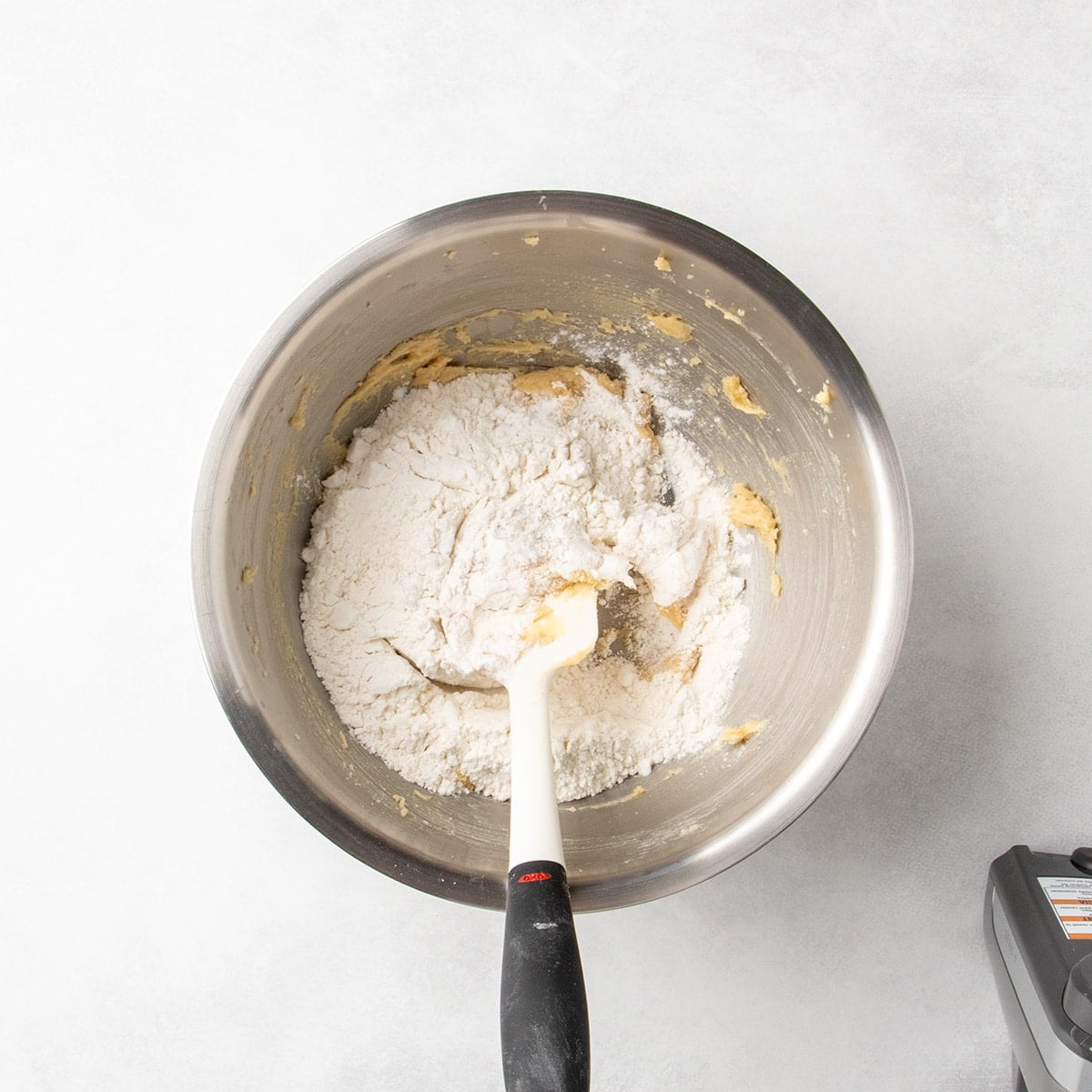 White flour added to a metal mixing bowl with creamed butter and sugar and a spatula in the bowl.
