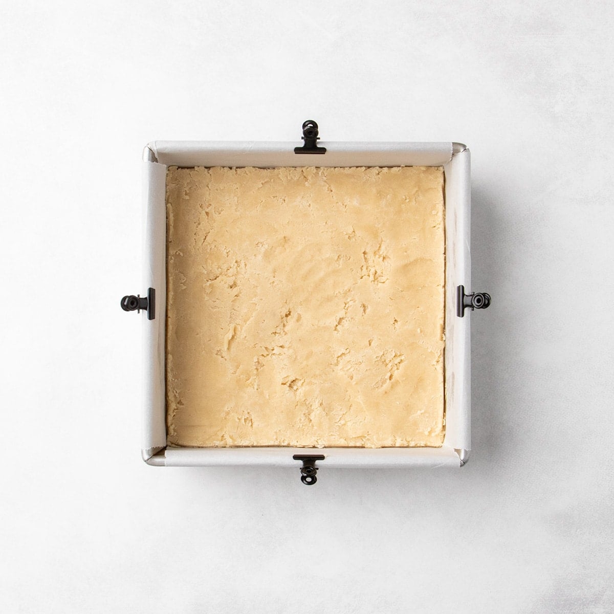 Raw light brown cookie dough pressed evenly into a parchment-lined square baking pan with binder clips around the edges.