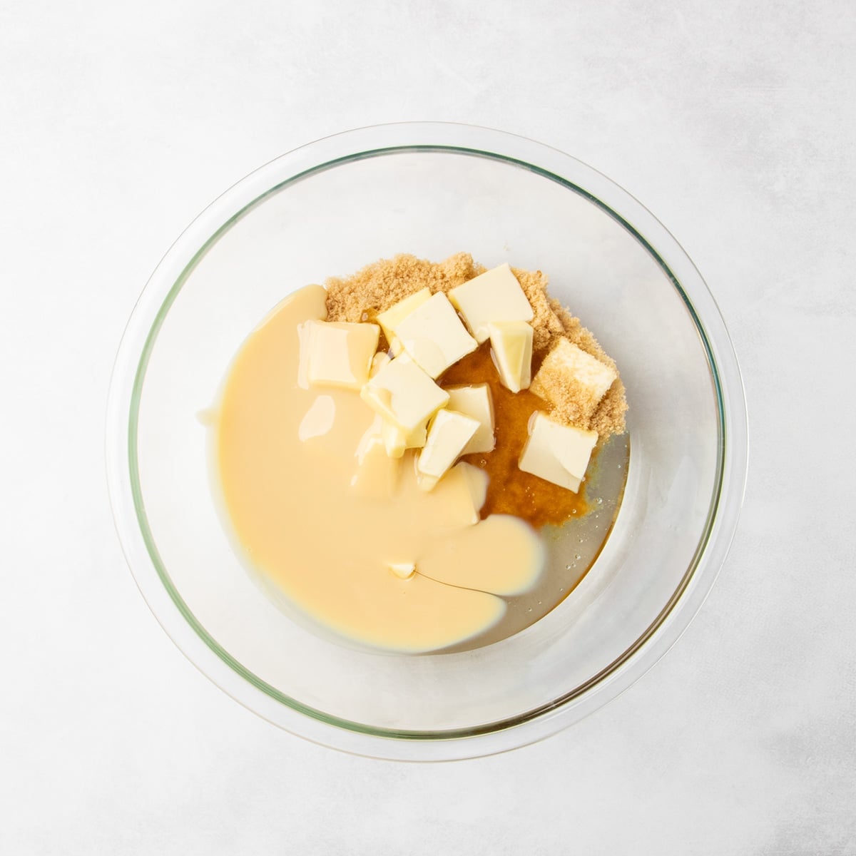 Brown sugar, pieces of butter, and sweetened condensed milk, and clear liquid ingredients in a clear glass mixing bowl.