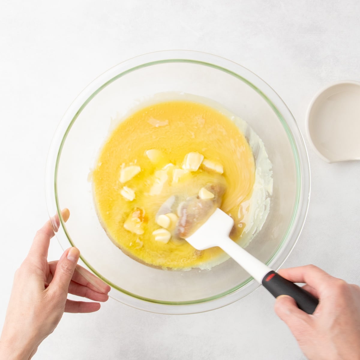Hands stirring melted butter and sugar mixture in a clear glass mixing bowl with a white spatula.