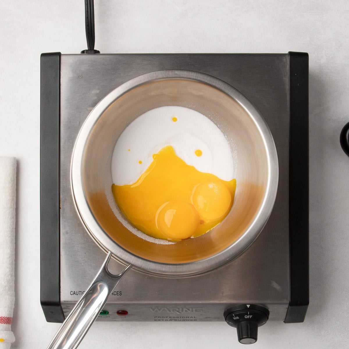 Metal saucepan on a hot plate with egg yolks and white granulated sugar inside.