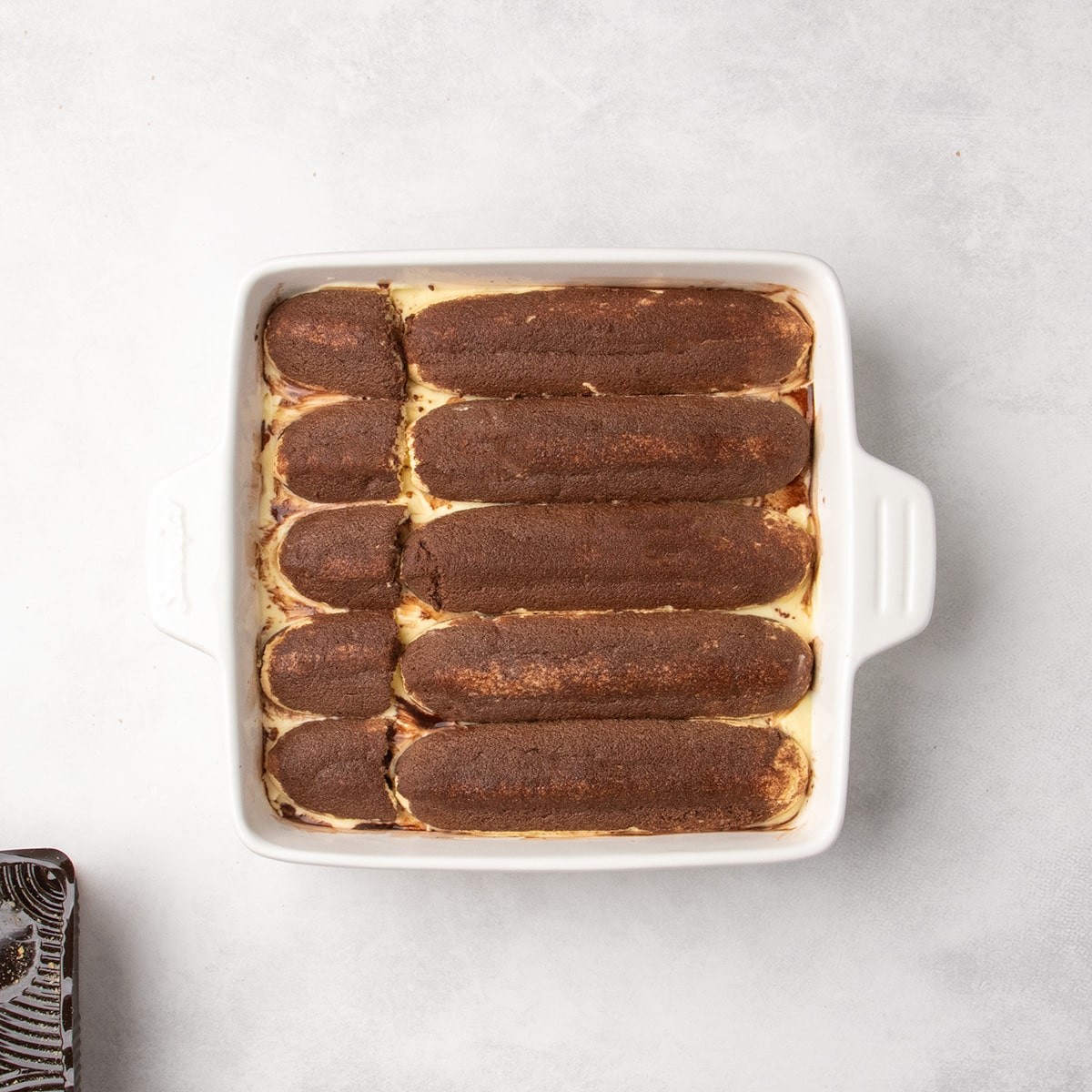 White baking dish with long cookies brushed with brown liquid arranged over a pale yellow layer.