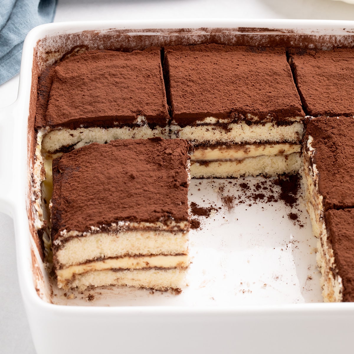 Sliced tiramisu in a white baking dish with one or more square pieces lifted out, showing pale layers and a dark powdered top.