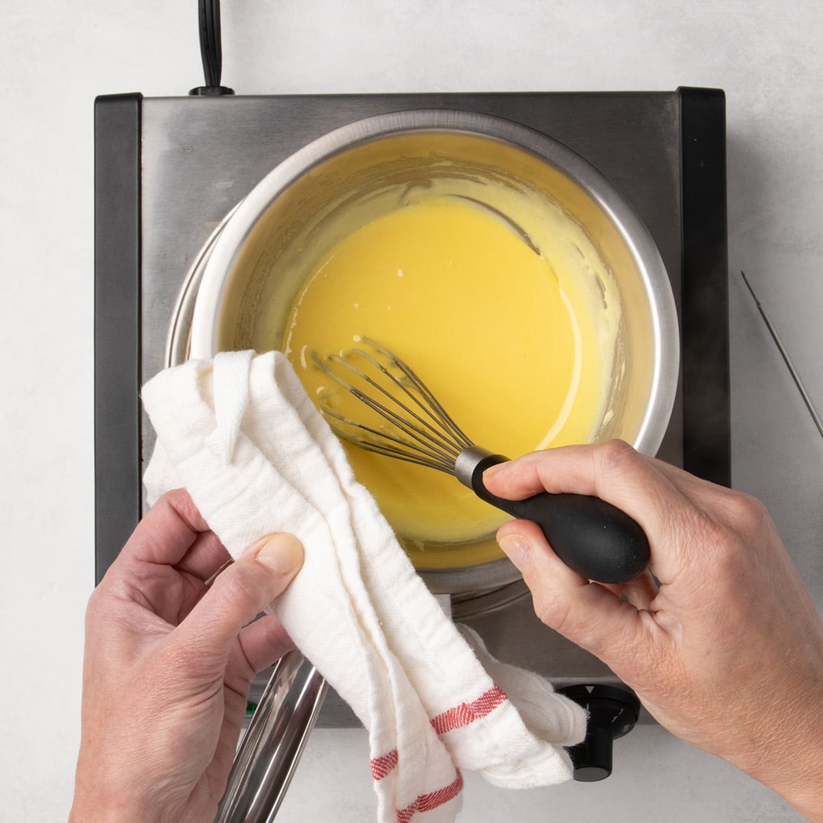 Hands holding a white tea towel on the edge of a small round metal mixing bowl and whisking a smooth yellow mixture.