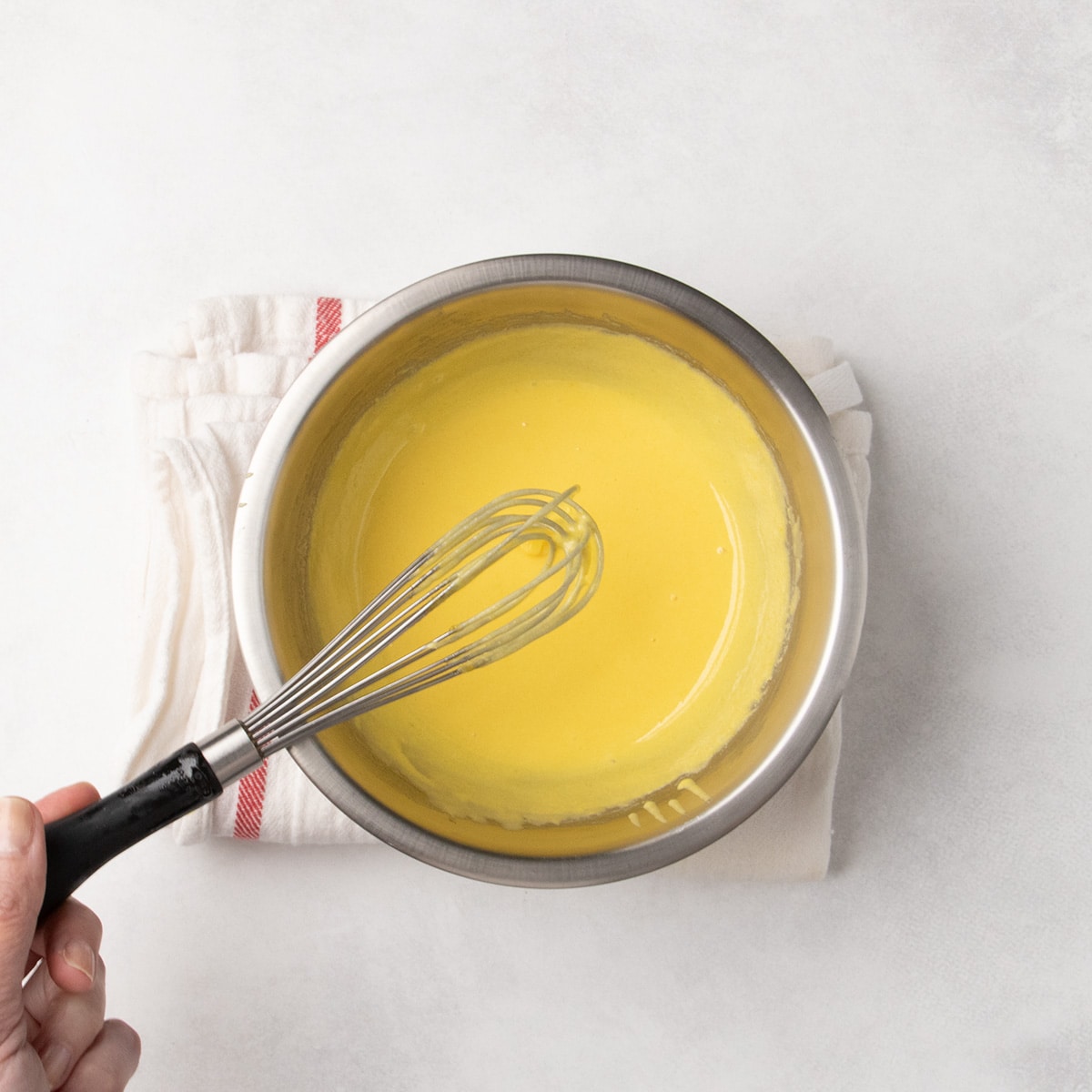 Metal round mixing bowl on a folded towel with a handheld whisk above a smooth yellow mixture in the bowl with a drizzle falling from the whisk into the bowl.