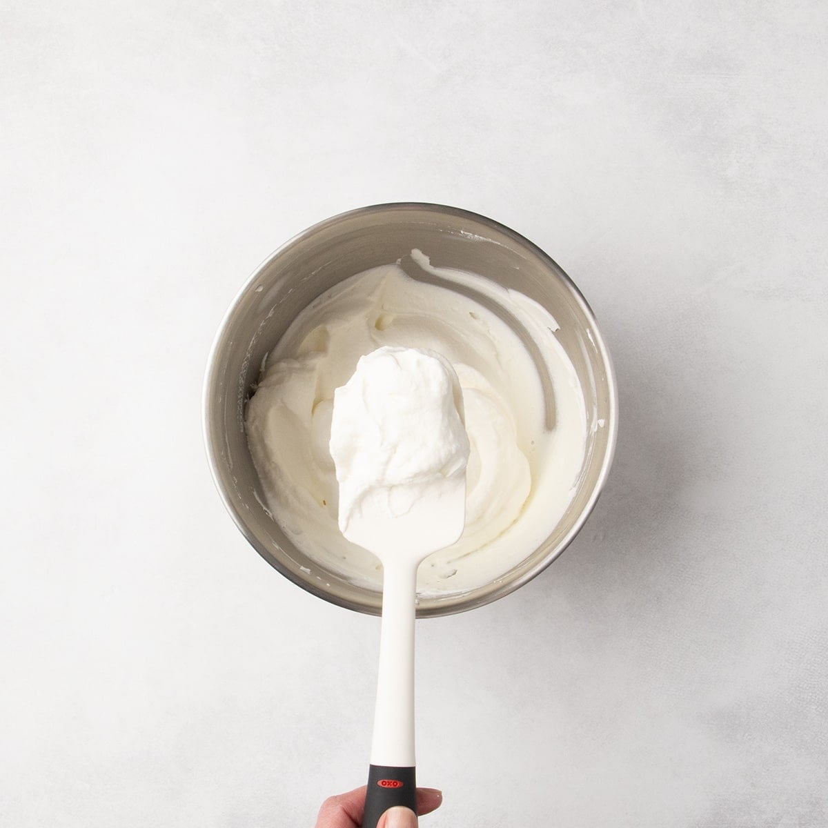 White spatula lifting fresh whipped cream from a metal mixing bowl.