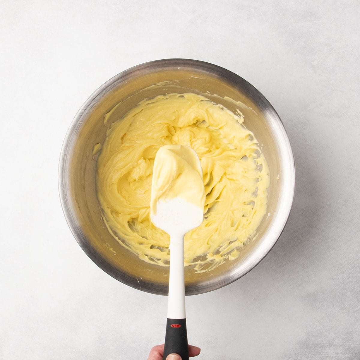 Thick pale yellow whipped mixture in a metal bowl with a white spatula held over the center.