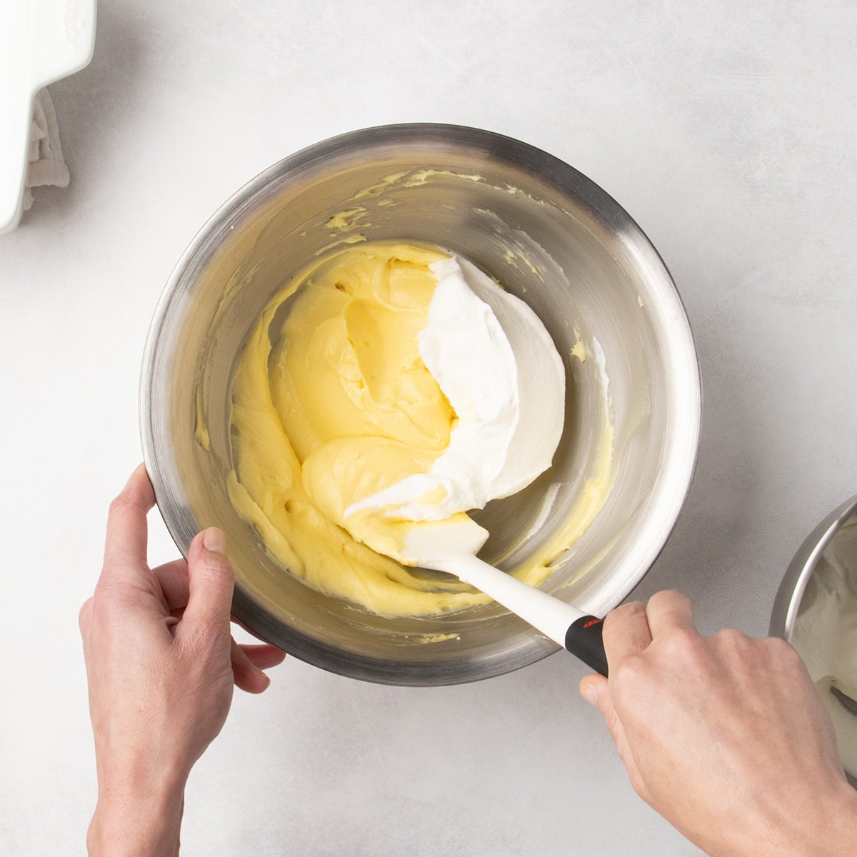 White spatula folding a white whipped cream mixture into a pale yellow mixture in a metal bowl.