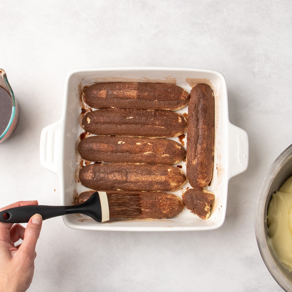 Long gluten free ladyfingers cookies lined up in a white baking dish being brushed with dark liquid.