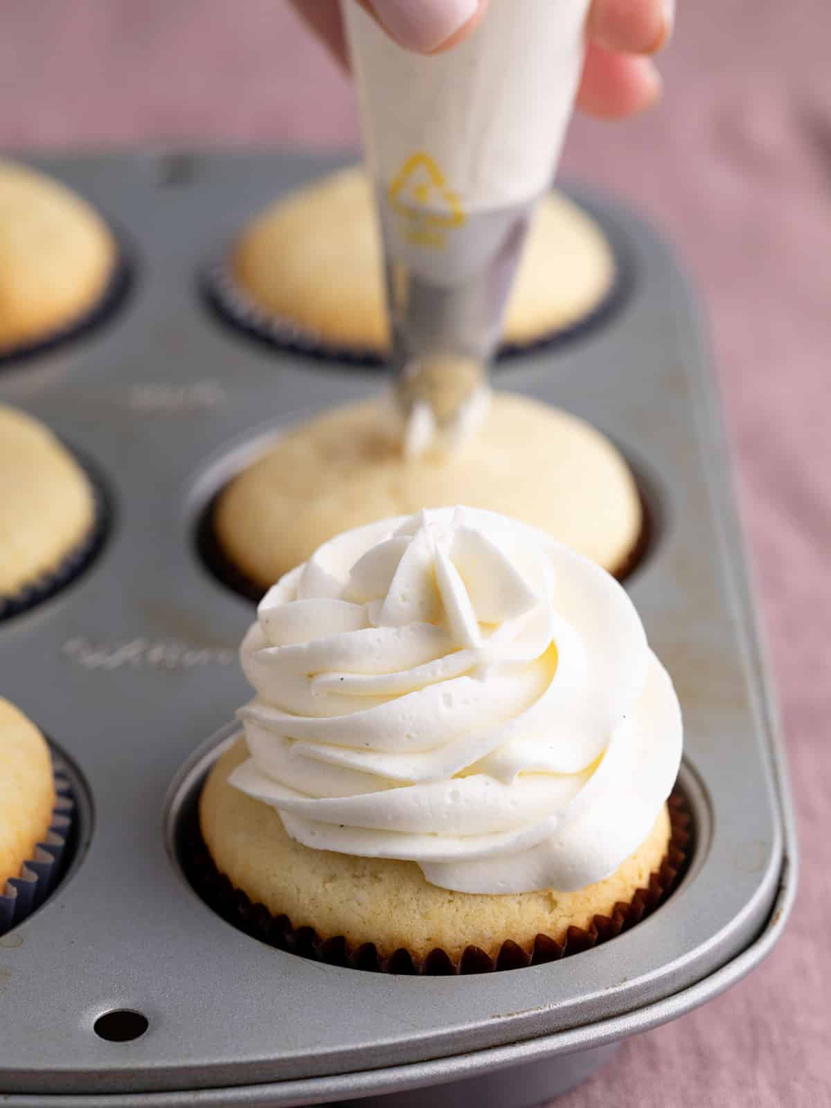 6 baked vanilla cupcakes in a muffin tin one with frosting swirl and hand holding piping bag on top of second cupcake in background