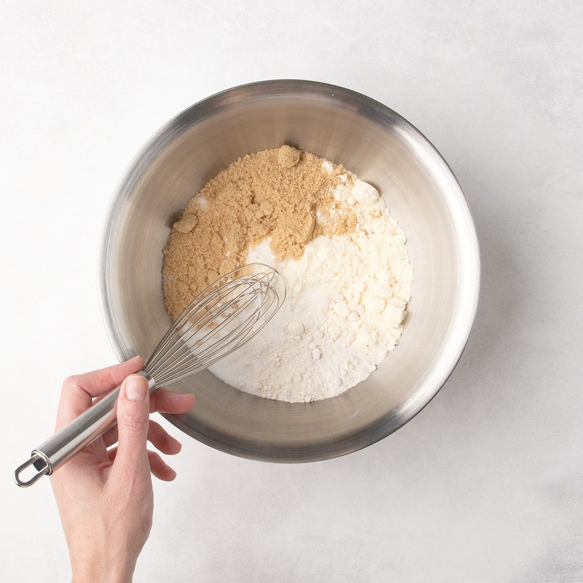 A metal mixing bowl with white and brown dry ingredients and a hand holding a whisk about to mix the contents of the bowl.