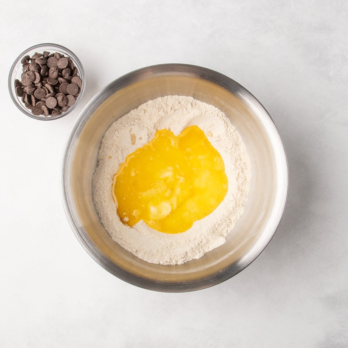 A metal mixing bowl with unmixed yellow liquid poured over dry ingredients, with a bowl of chocolate chips nearby.