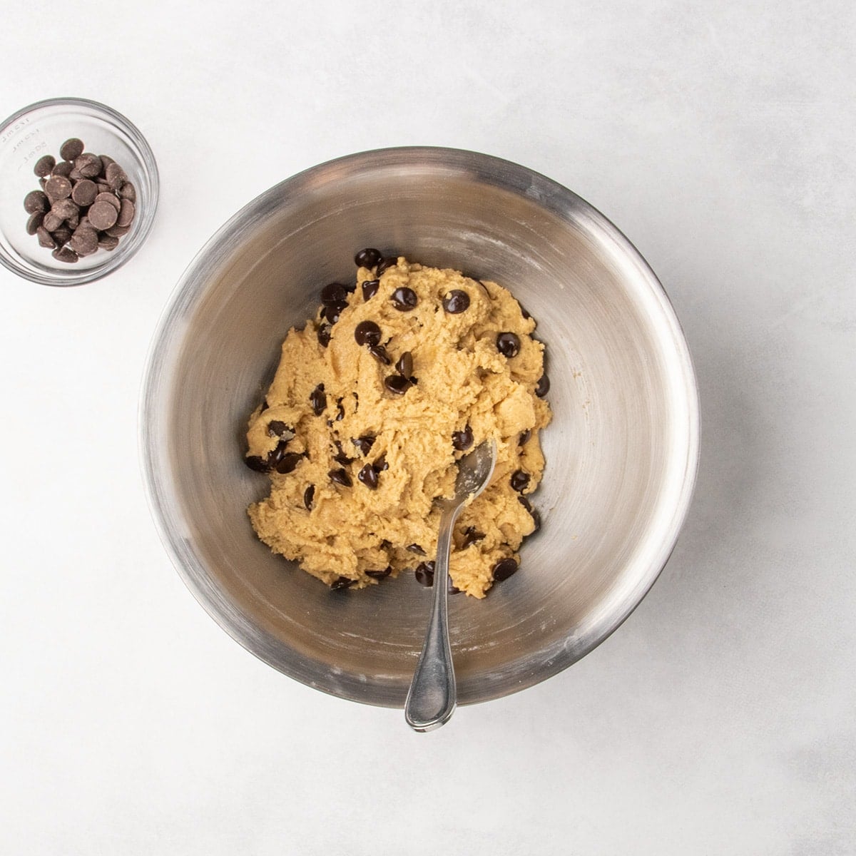 A metal mixing bowl with chocolate chips mixed into pale tan cookie dough and a spoon inside.