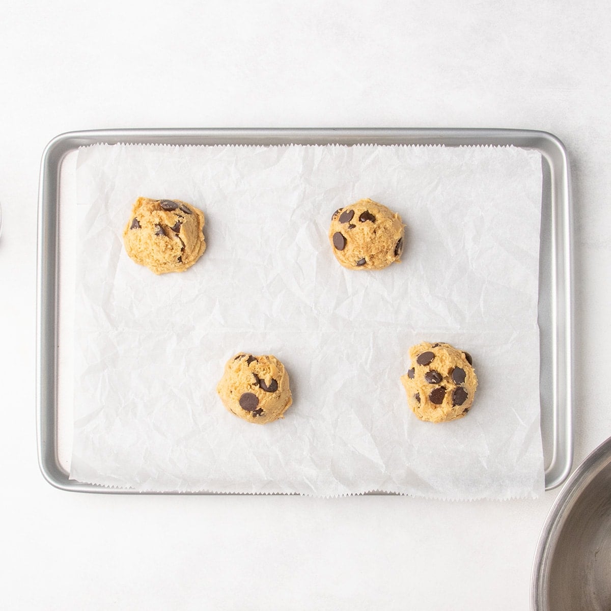 Four scoops of chocolate chip cookie dough on a parchment-lined baking sheet.