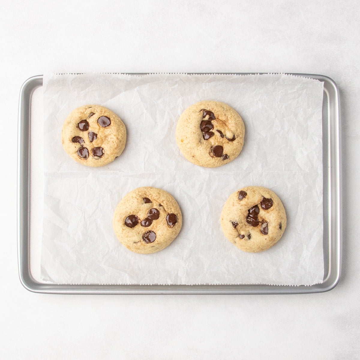 Four baked pale chocolate chip cookies on a parchment-lined baking sheet.