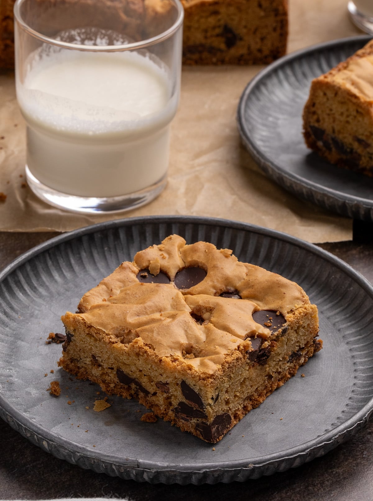 Chocolate chip cookie bar on a small dark gray round plate with a glass of milk and another plated cookie bar in the background.