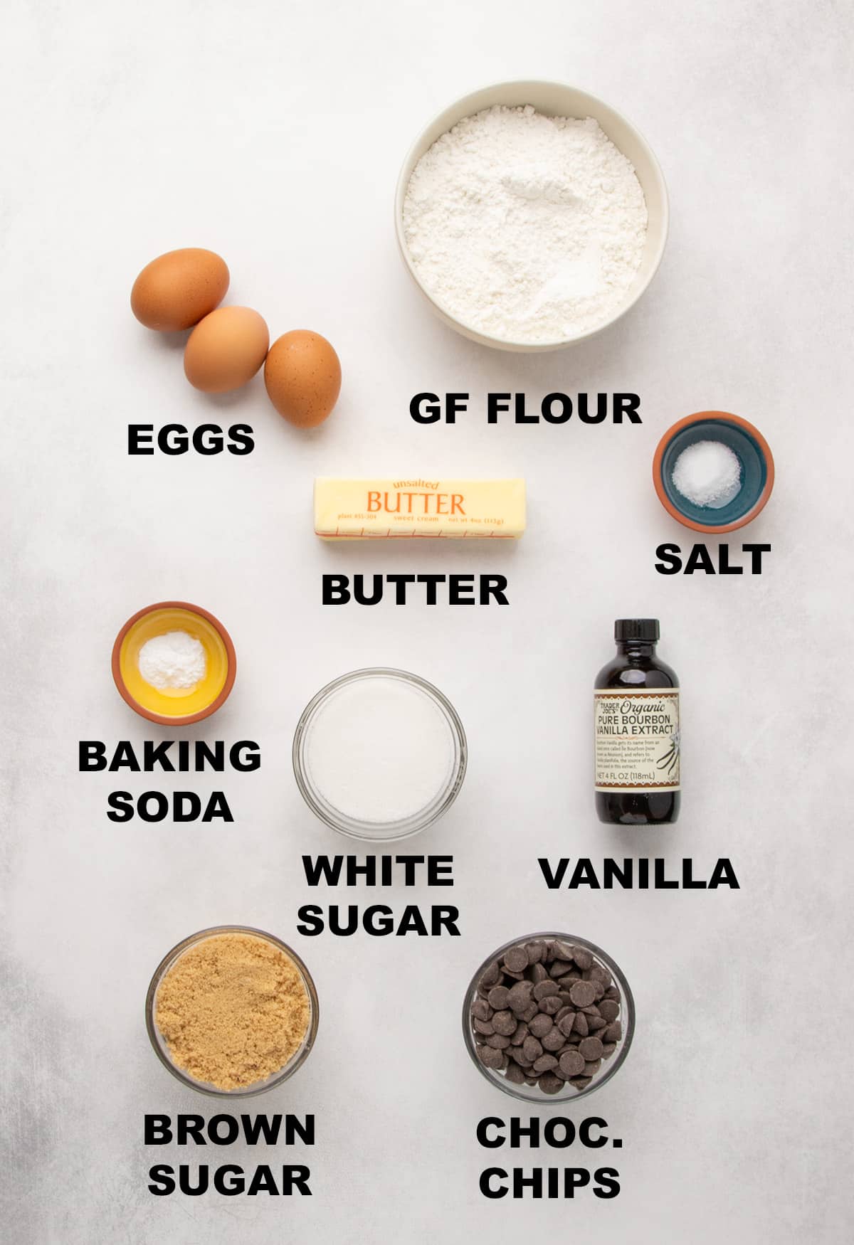 Overhead view of eggs, GF flour mixture, salt, butter, baking soda, white sugar, vanilla, light brown sugar, and chocolate chips labeled on a light surface.