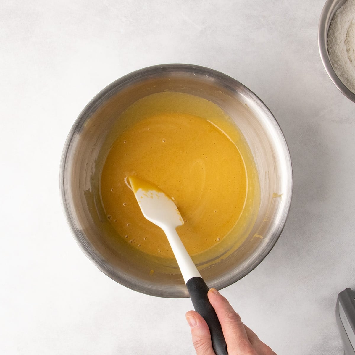 Overhead view of a smooth yellow mixture in a metal mixing bowl with hand holding a white spatula.