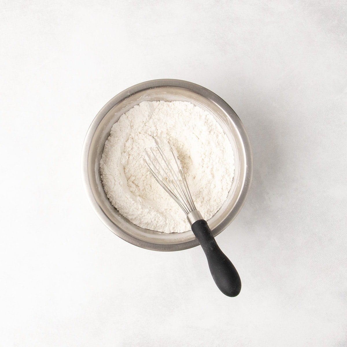 Overhead view of white flour mixture in a small metal bowl with a small wire whisk with a black handle.