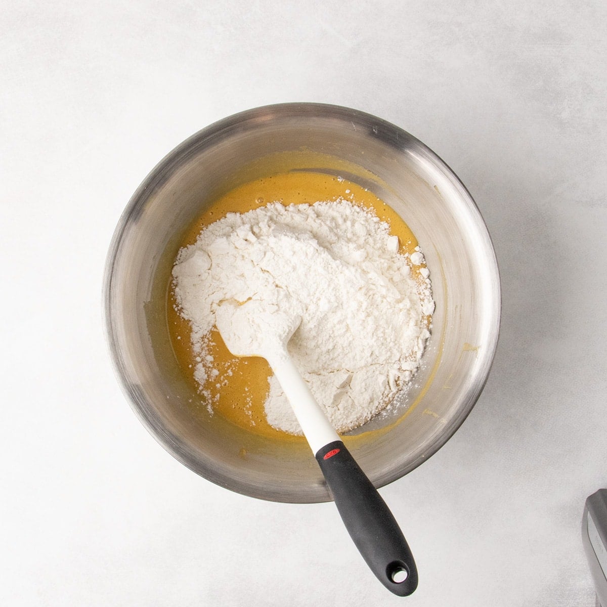 Overhead view of white flour mixture on top of yellow batter in a metal mixing bowl with a white spatula.