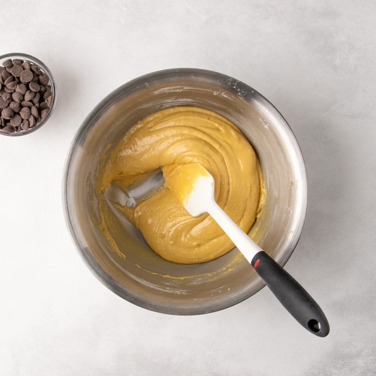 Overhead view of thick yellow batter in a metal mixing bowl with a white spatula and a small bowl of chocolate chips nearby.