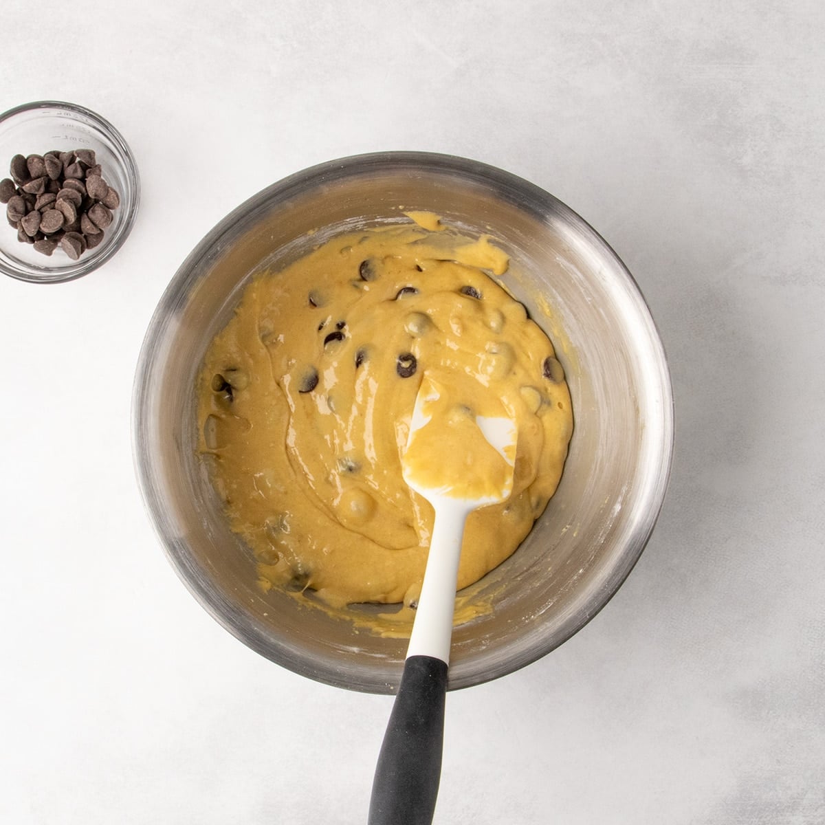 Overhead view of chocolate chips mixed into thick yellow batter in a metal mixing bowl with a white spatula.
