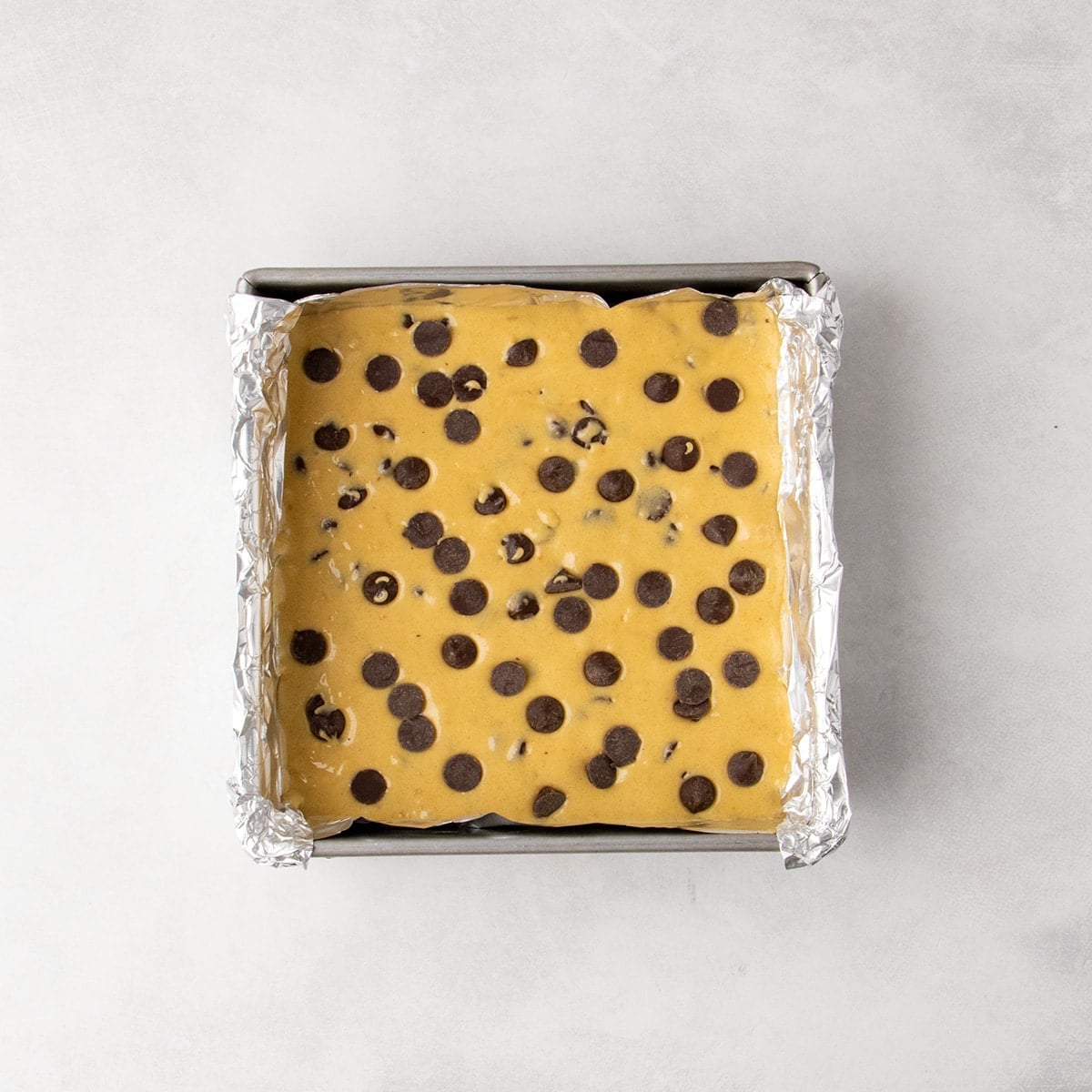 Overhead view of yellow batter with chocolate chips spread in a foil-lined square baking pan.