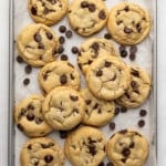 Close-up of baked and lightly golden brown gluten free dairy free chocolate chip cookies in a pile with loose chocolate chips on a parchment-lined baking sheet.