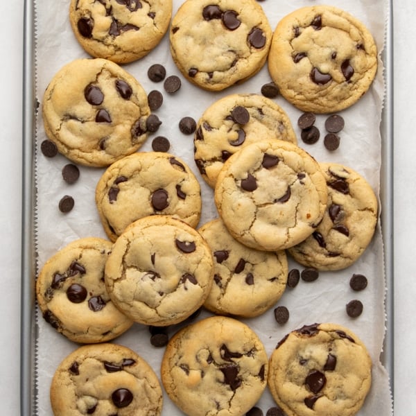 Close-up of baked and lightly golden brown gluten free dairy free chocolate chip cookies in a pile with loose chocolate chips on a parchment-lined baking sheet.