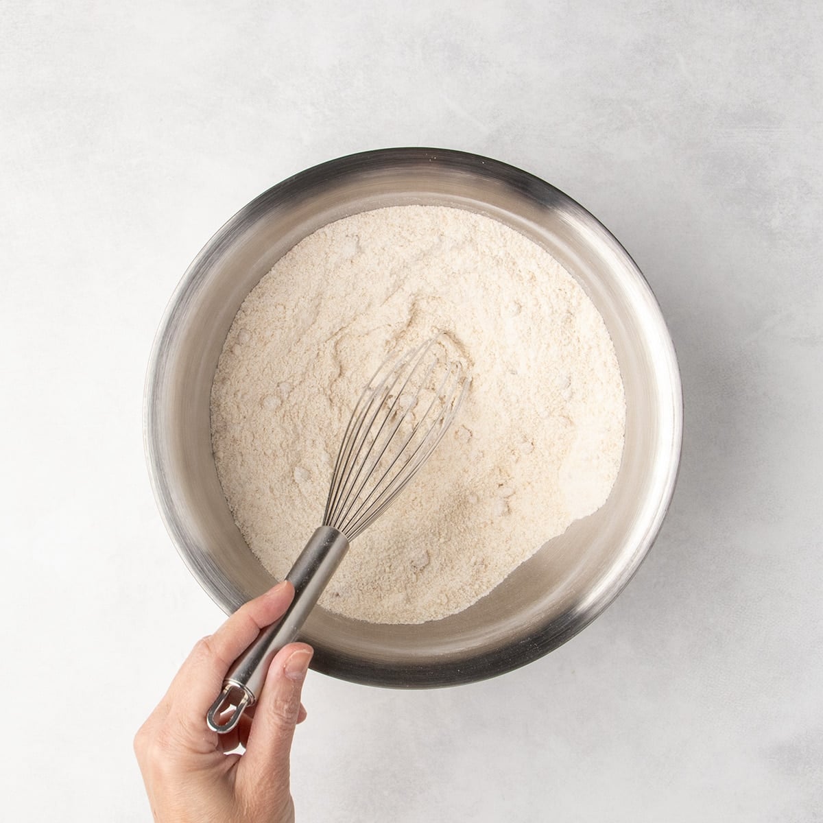 Hand holding a whisk in a metal bowl of combined light tan dry ingredients.