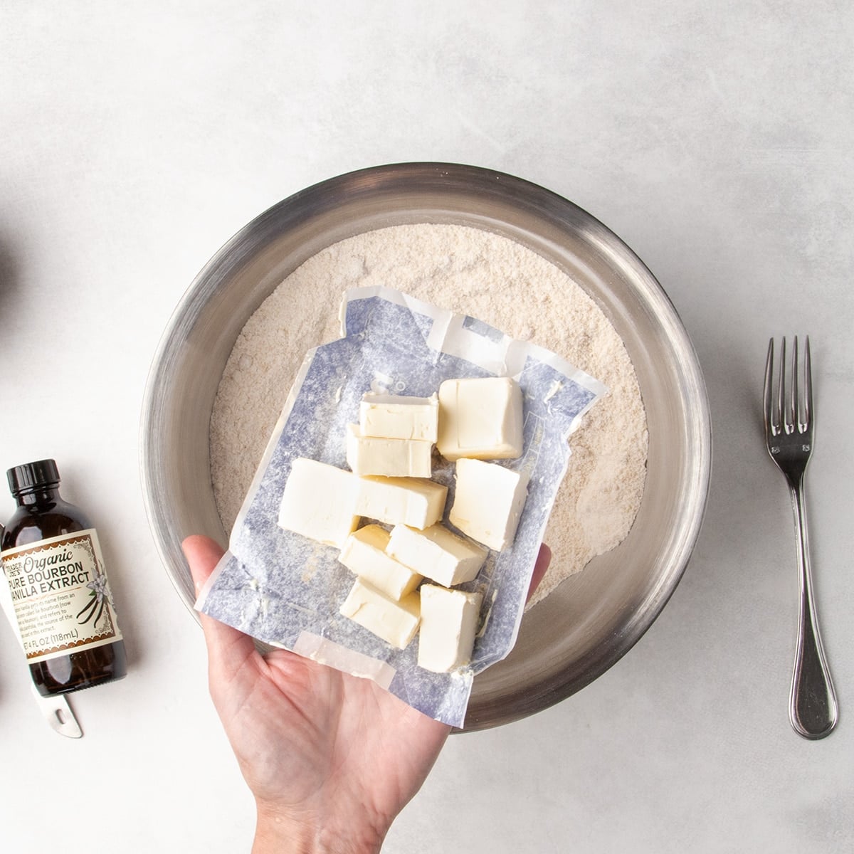 Hand holding wrapper with cubed vegan butter above a metal bowl of dry ingredients, with a vanilla bottle and fork nearby.