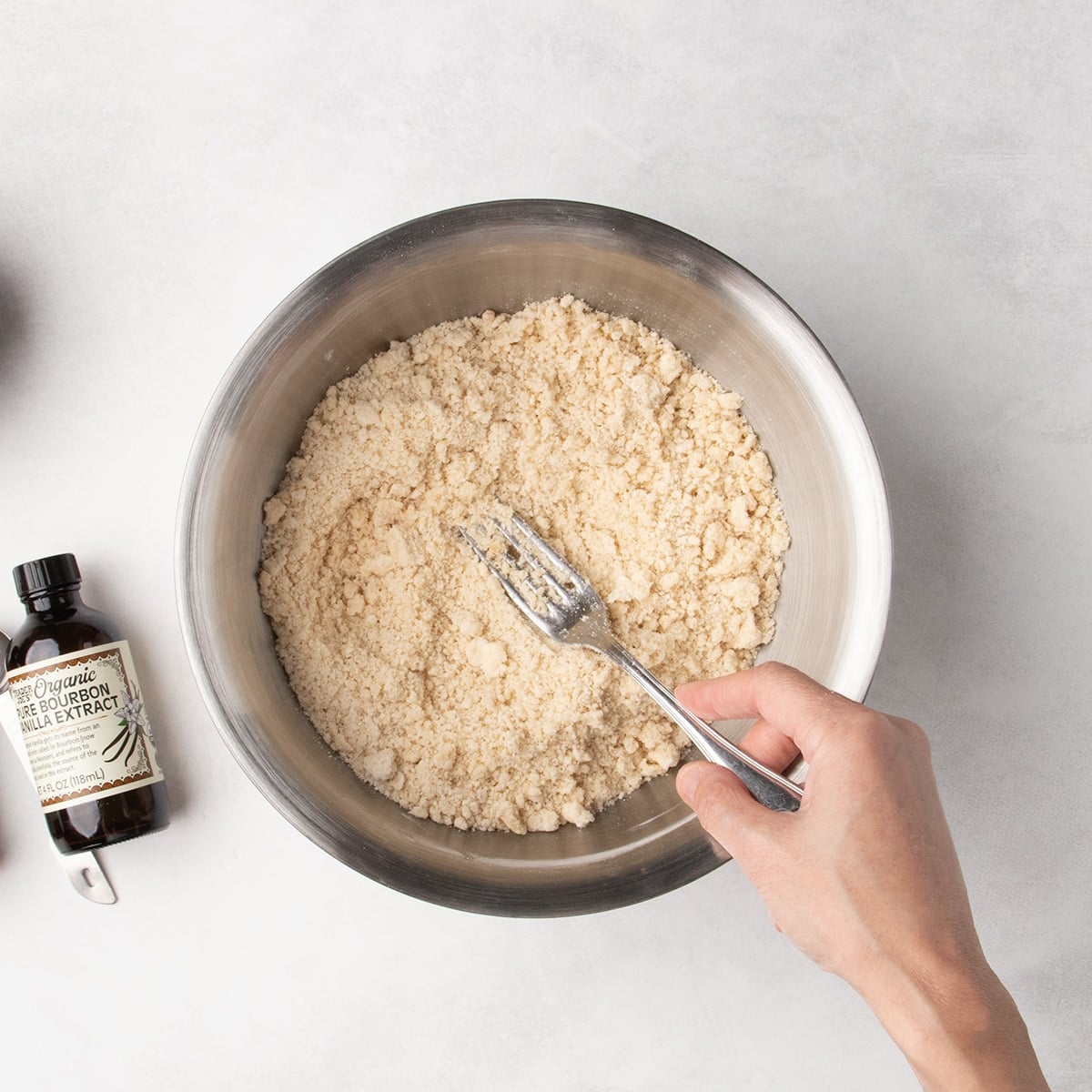 Fork mixing moist light tan crumbles in a metal bowl, with a vanilla bottle nearby.