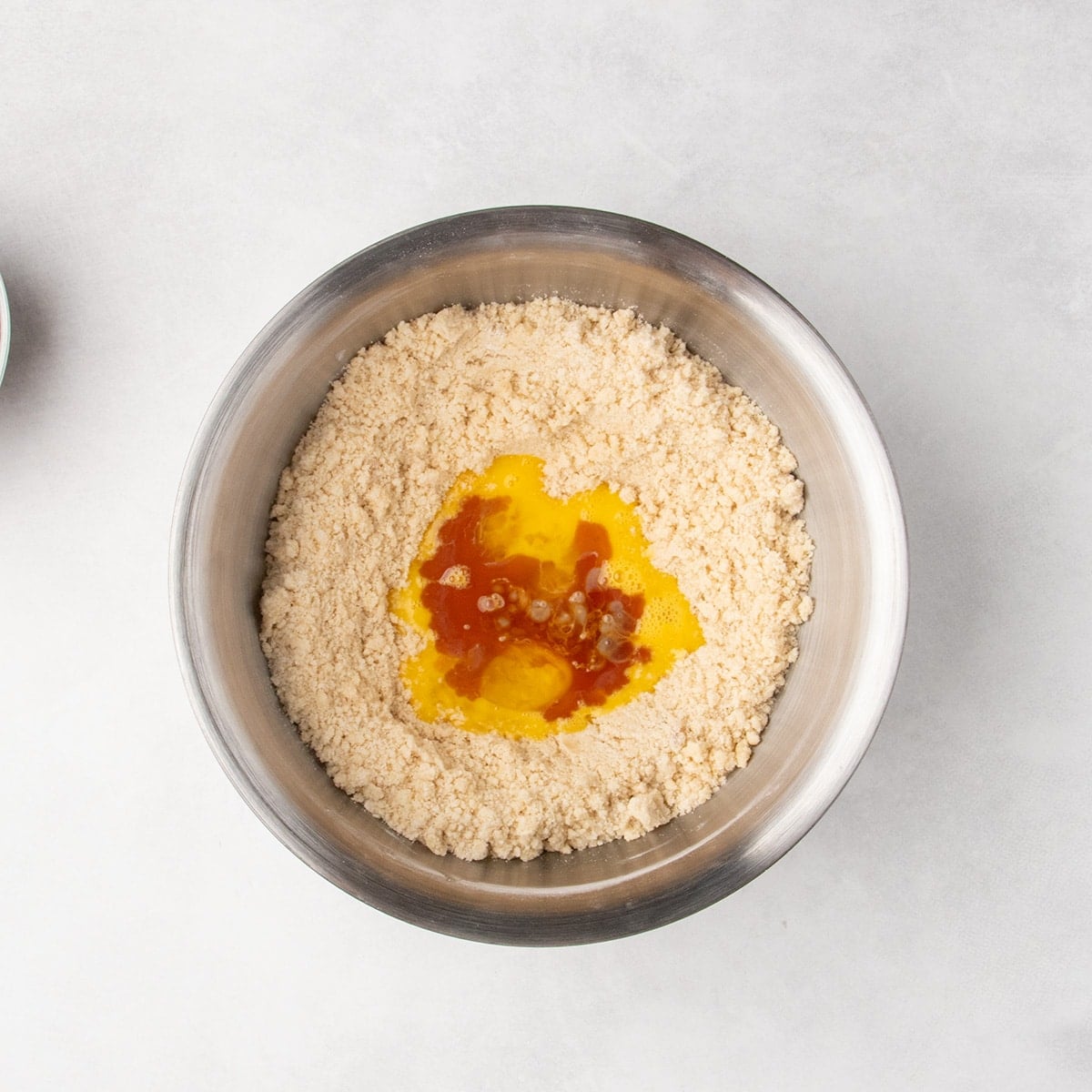 Metal bowl with light tan dough crumbles on bottom, and a well filled with beaten eggs and vanilla.
